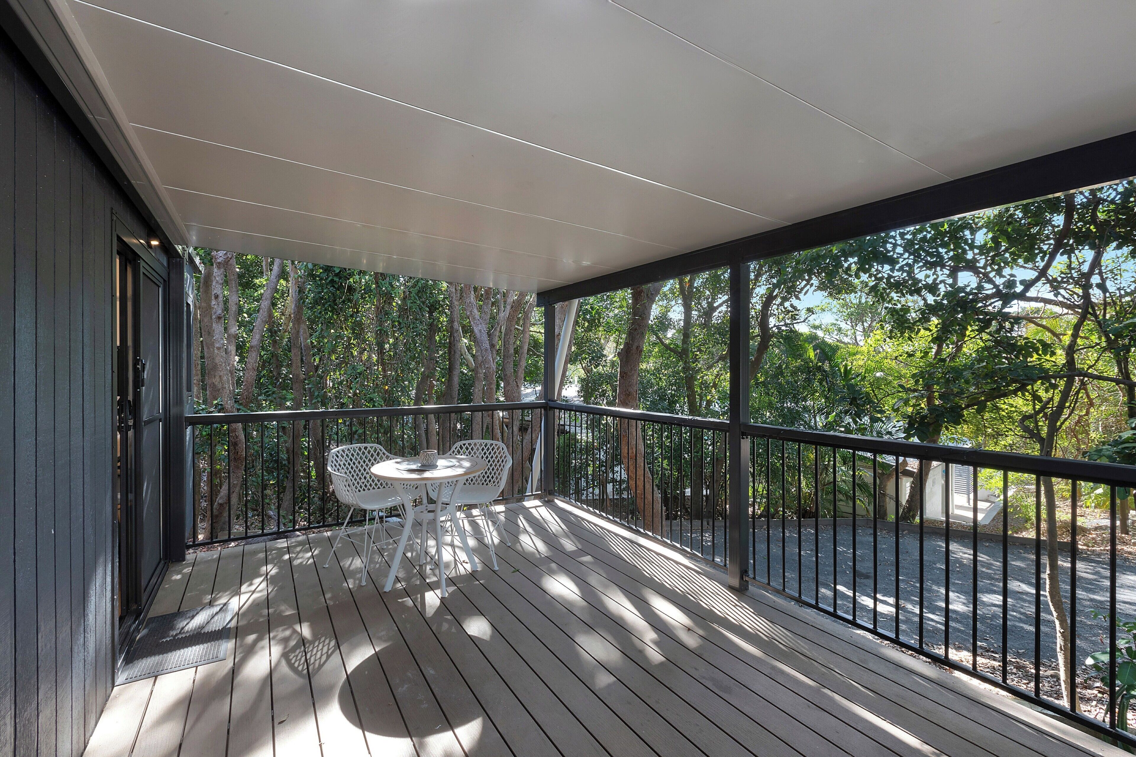 A wooden deck with a table and chairs, surrounded by a railing, overlooking a road and trees.