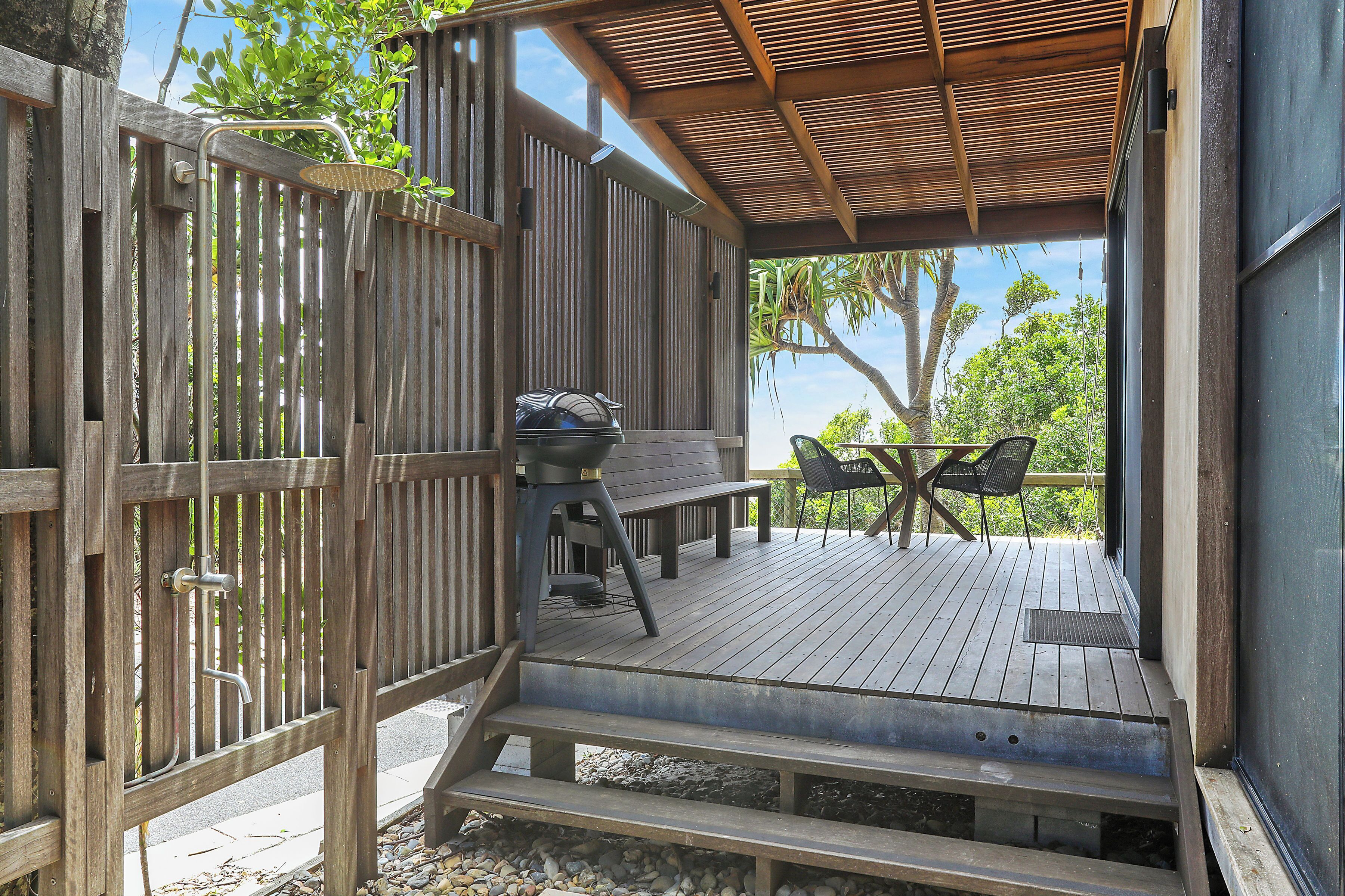 Una terraza de madera con zona de estar cubierta, una parrilla y vistas a árboles.