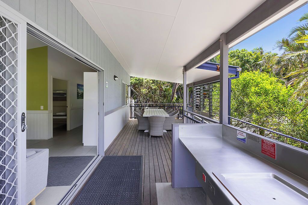 A covered outdoor area with a wooden deck, a dining table with chairs, and a view of lush greenery.