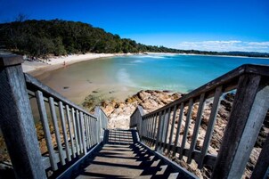 Una escalera de madera que lleva a una playa con aguas azul turquesa y una ladera boscosa al fondo.