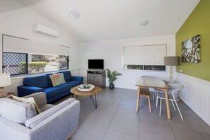 A modern living room with a blue sofa, a grey armchair, a wooden coffee table, and a dining area with a table and chairs. - Reflections Byron Bay - Holiday Park (Byron Bay)