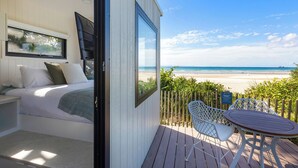 A bedroom with a large window overlooking a beach, a small balcony with a table and chairs, and a bed with pillows. - Reflections Byron Bay - Holiday Park (Byron Bay)