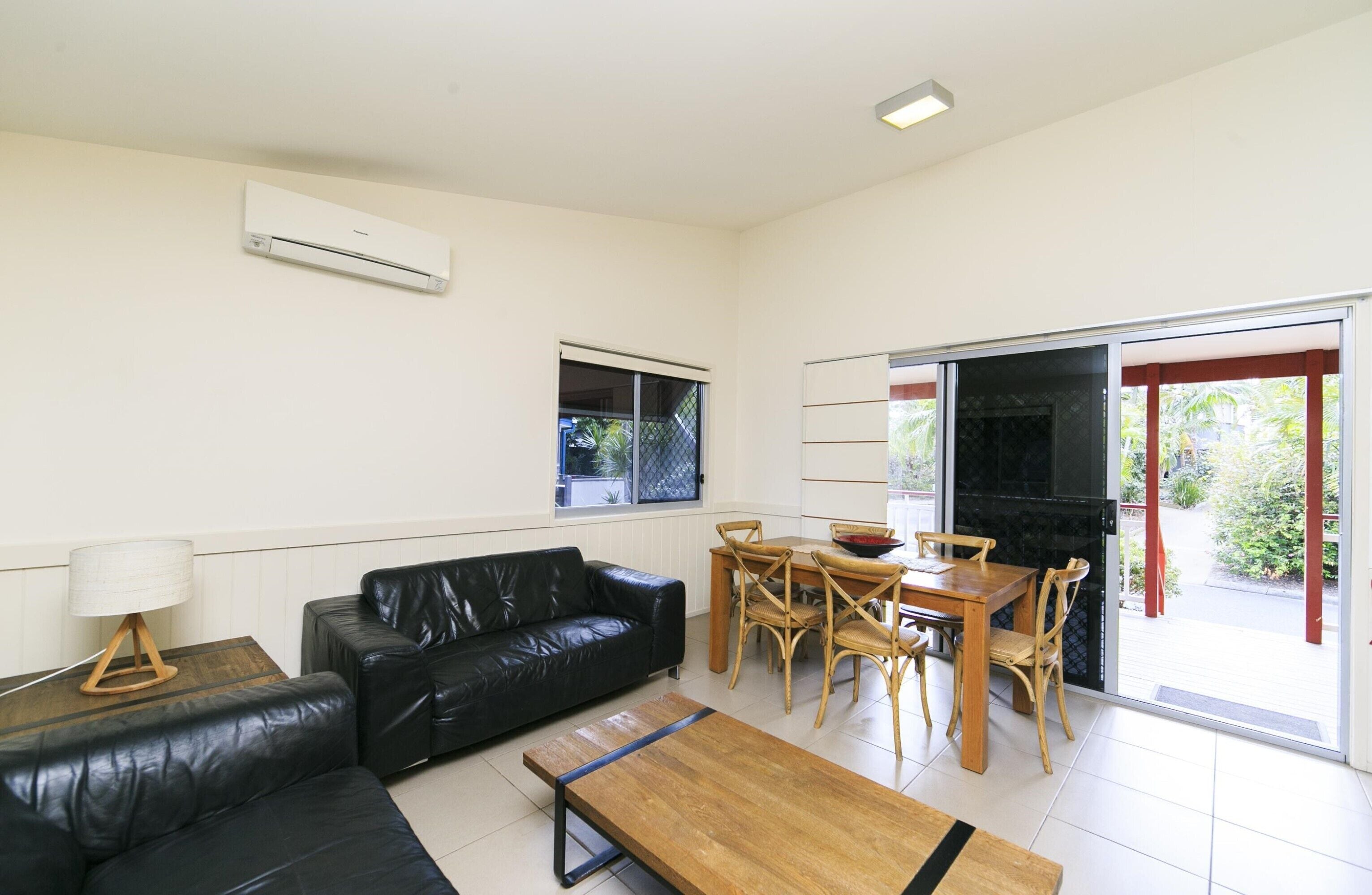 Una sala de estar moderna con un sofá de cuero negro, una mesa de centro de madera y un comedor con sillas y mesa de madera. Hay una puerta corrediza de vidrio que da a un balcón con una parrilla negra.