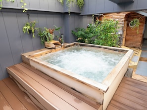 Tinaja de madera con escalones que la conducen, rodeada de macetas con plantas y una estructura de cabaña de madera.