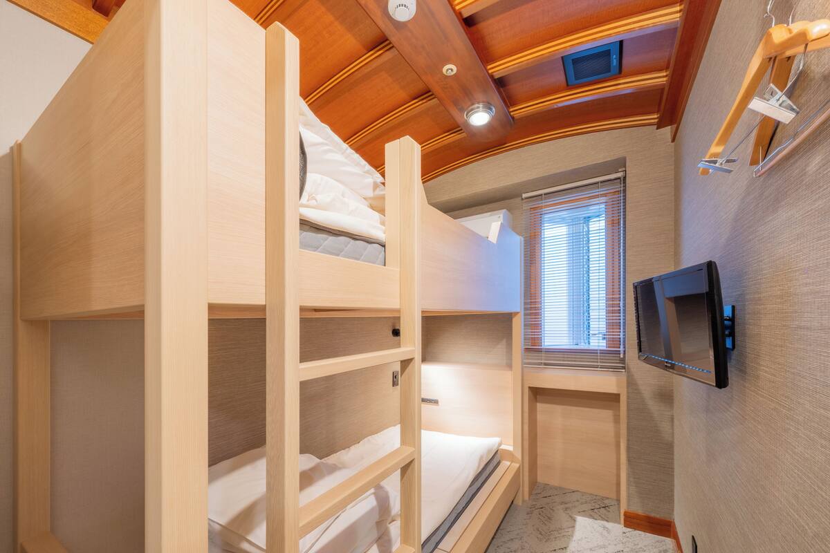 A bunk bed room with a television on the wall, a window with blinds, and a ceiling vent.