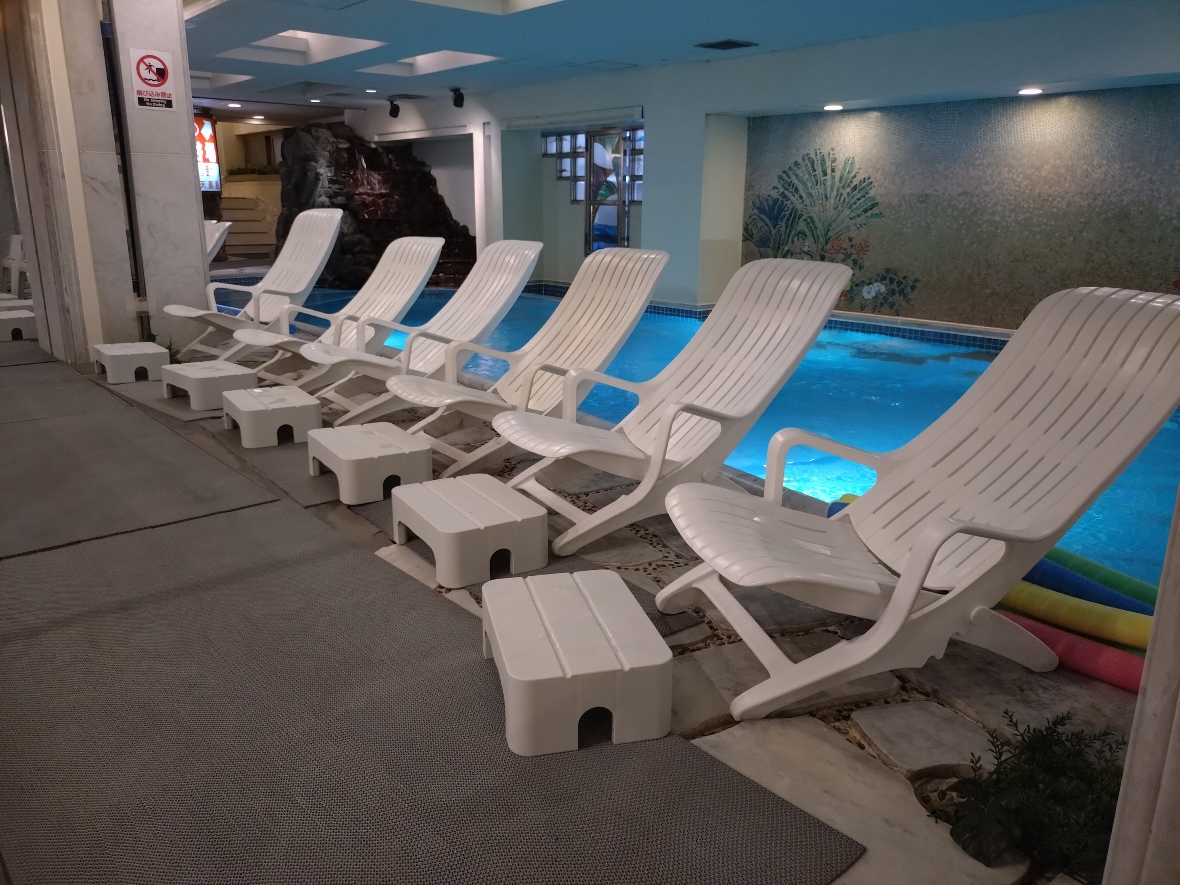 Un área junto a la piscina con sillones blancos y pequeños taburetes, piso de baldosas y un mural en la pared.