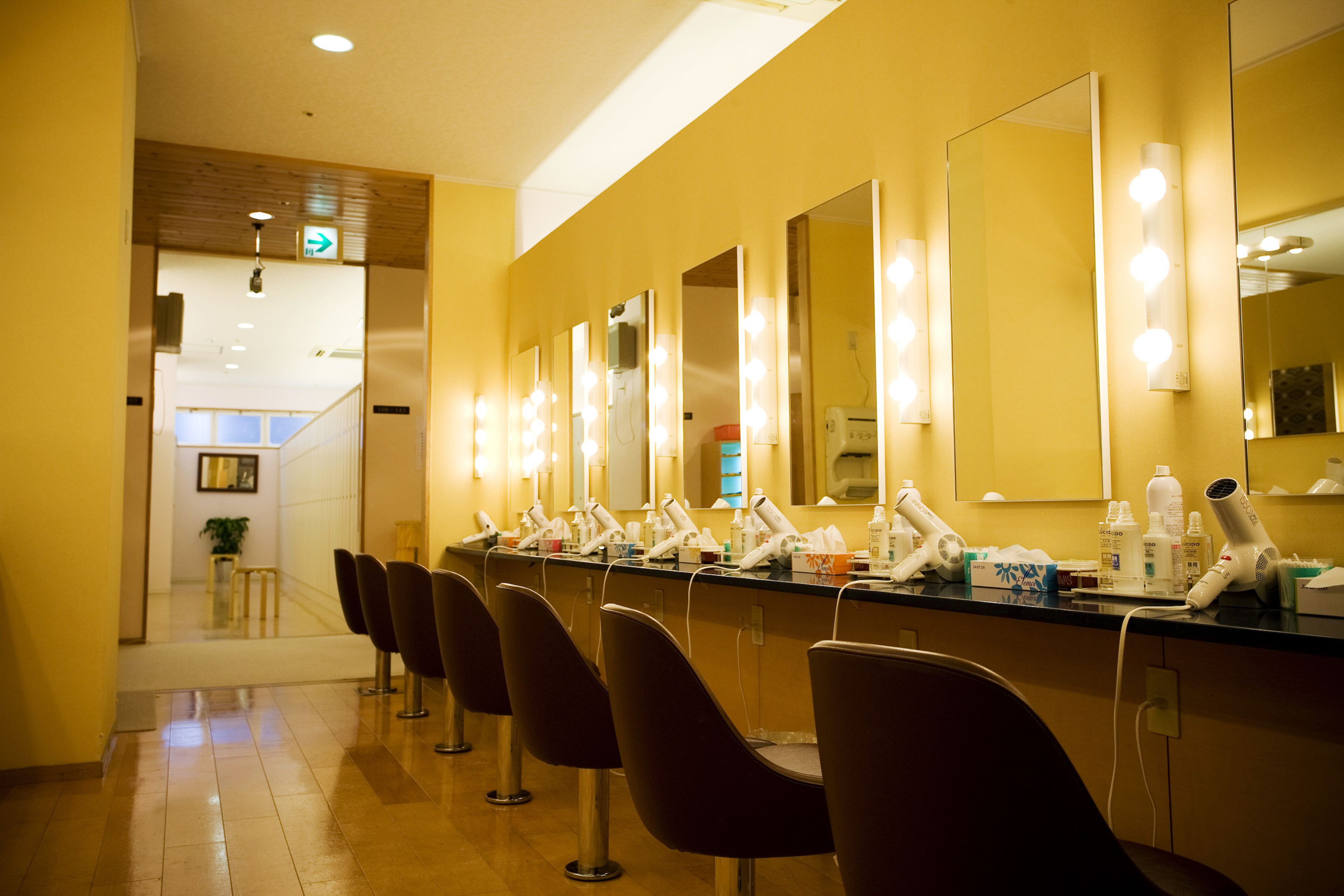 A row of salon chairs with mirrors and lighting, a counter with hairdryers, and a staircase in the background.
