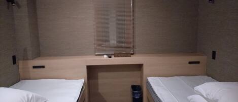 A small, compact room with two single beds, a window with blinds, a built-in desk, and a trash bin.