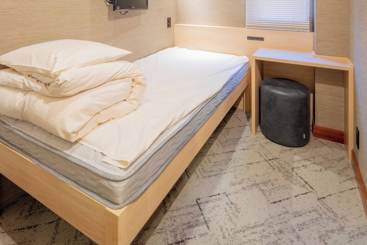 A single bed with a mattress, pillows, a bedside table, a small stool, a wall-mounted TV, and an air conditioning unit.