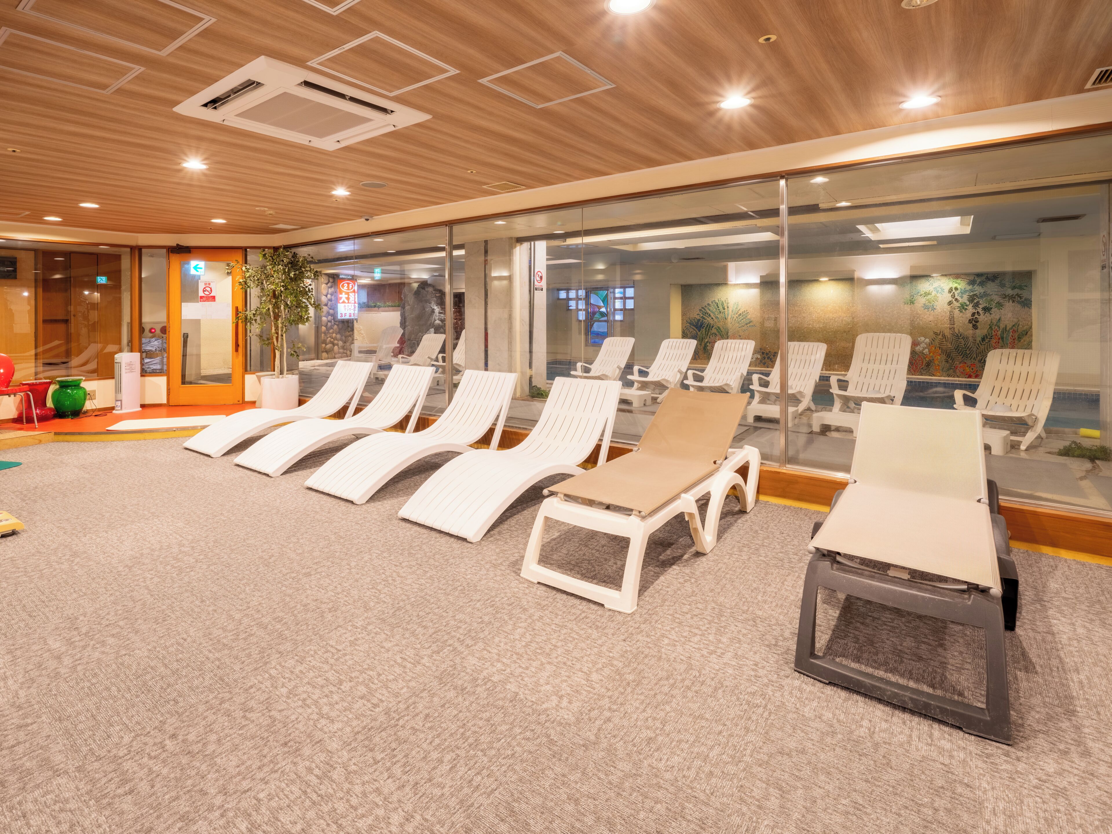 A room with white lounge chairs, a carpeted floor, wooden ceiling panels, and a glass wall with a view.
