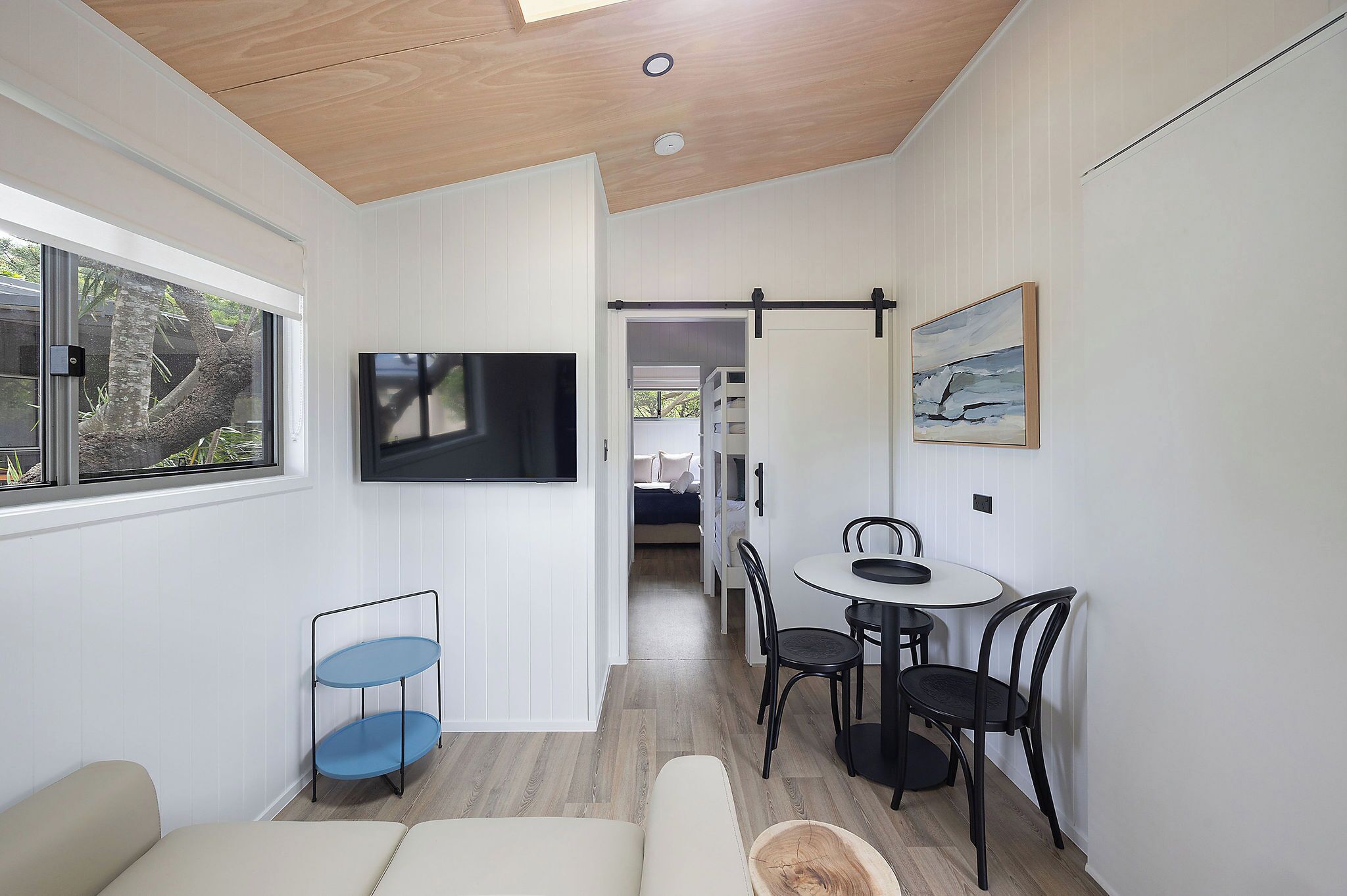 A modern, compact living space with a dining area, a TV mounted on the wall, and a view of the outdoors through a window.