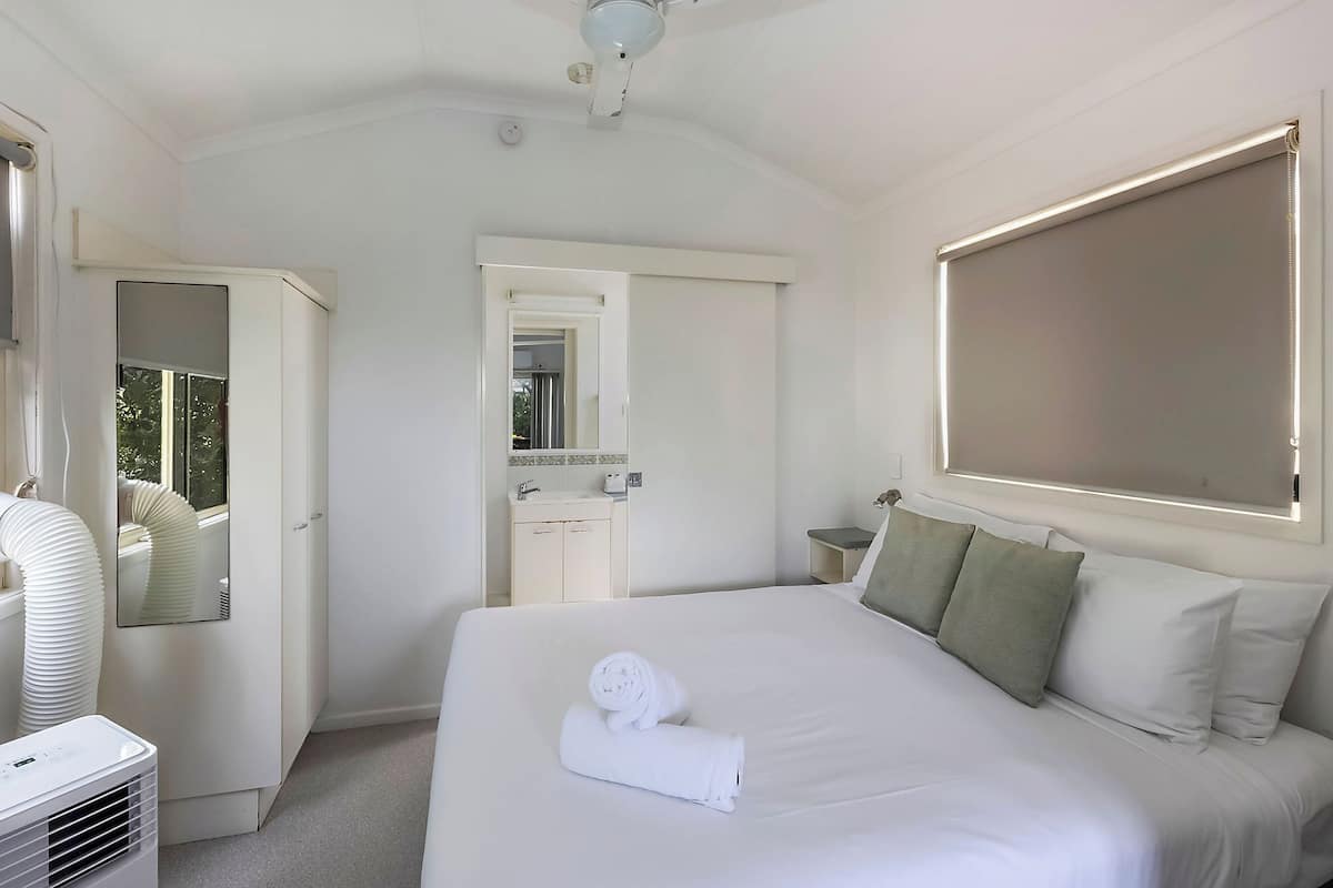 Quarto de hotel com uma cama grande, uma mesa de cabeceira, um espelho, uma janela com persianas e um ventilador de teto.