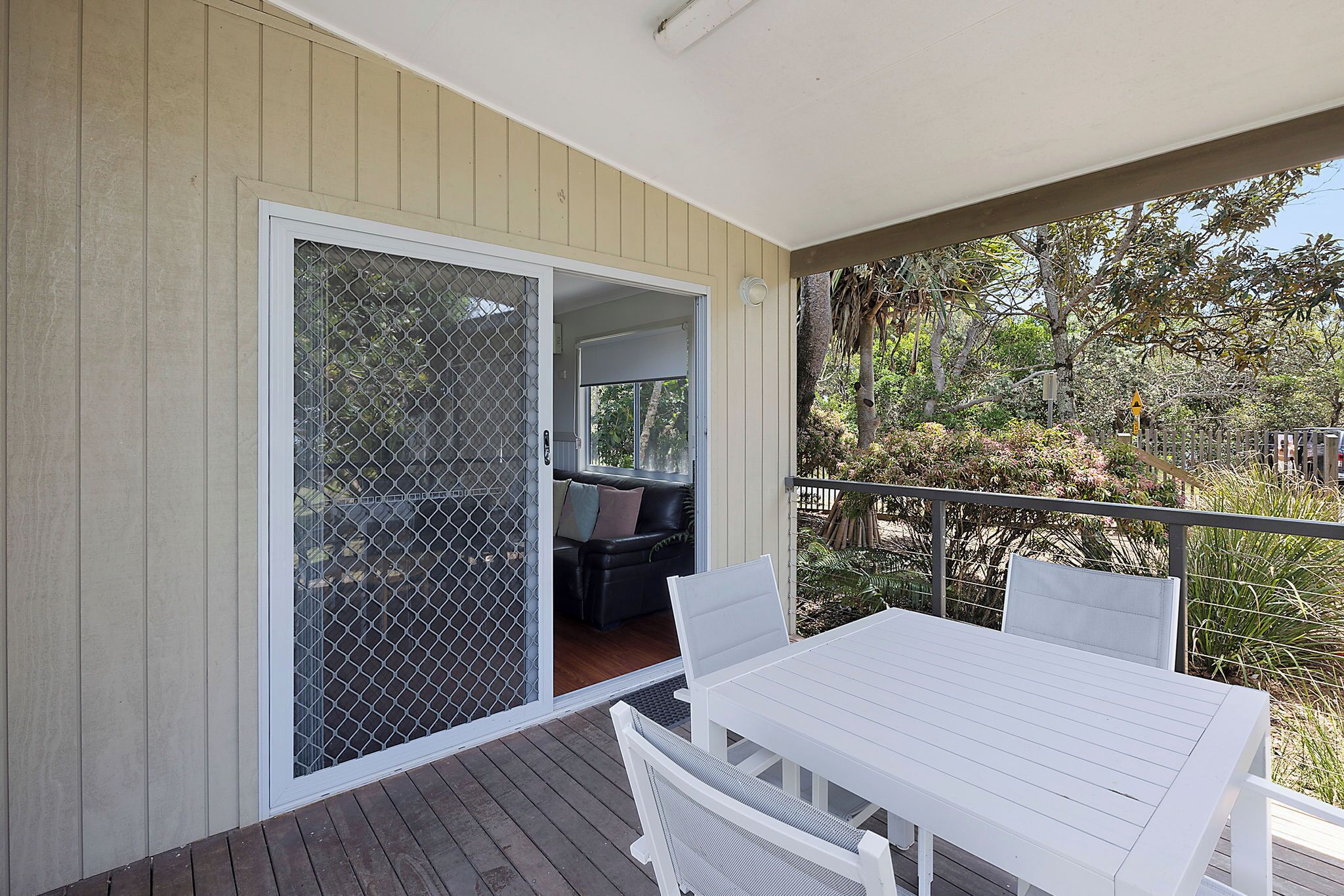 A balcony with a white table and chairs, a sliding glass door leading to a room with a black leather sofa, and a view of a garden with trees and a fence.