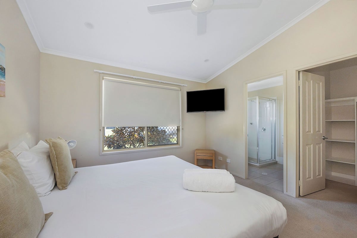 Quarto com uma cama grande, um ventilador de teto, uma TV na parede e janela com vista para árvores.