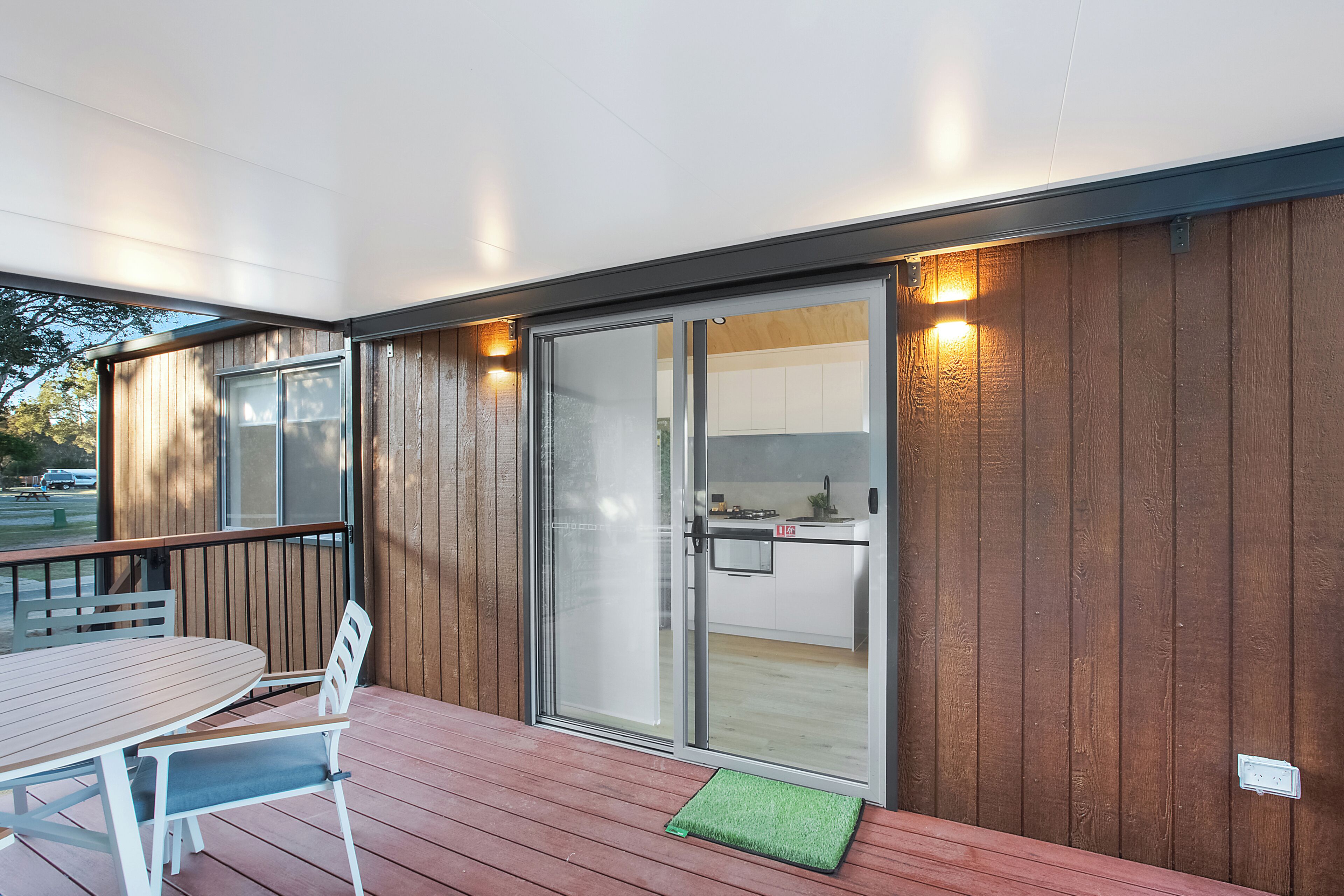 Una terraza de madera con una mesa y sillas, una puerta corrediza de vidrio que da a una cocina y un tapete verde pequeño en la terraza.