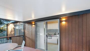 Una terraza de madera con una mesa y sillas, una puerta corrediza de vidrio que da a una cocina y un tapete verde pequeño en la terraza.