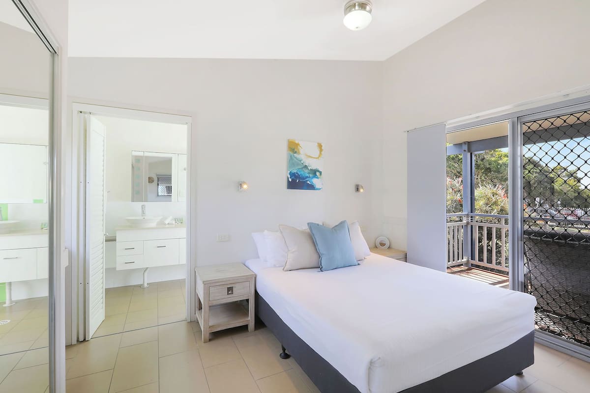 Quarto com cama, mesa de cabeceira, espelho, pintura e varanda com vista.