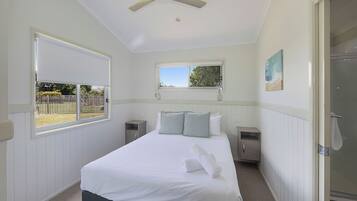 Un dormitorio con cama, ventilador de techo, ventana, un cuadro y la puerta del baño.