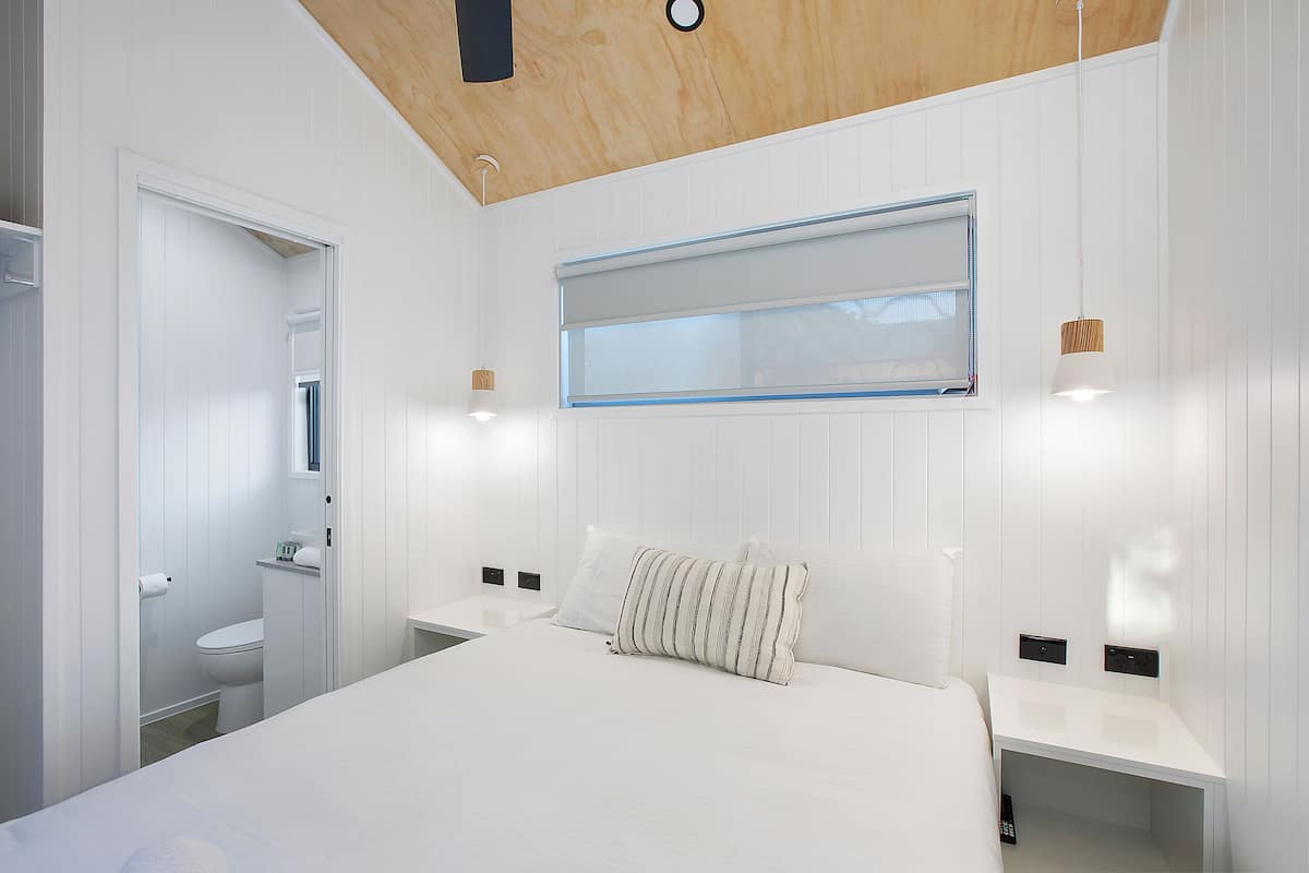 Quarto moderno com uma cama grande, mesa de cabeceira, estante e janela com persianas, além de teto de madeira.