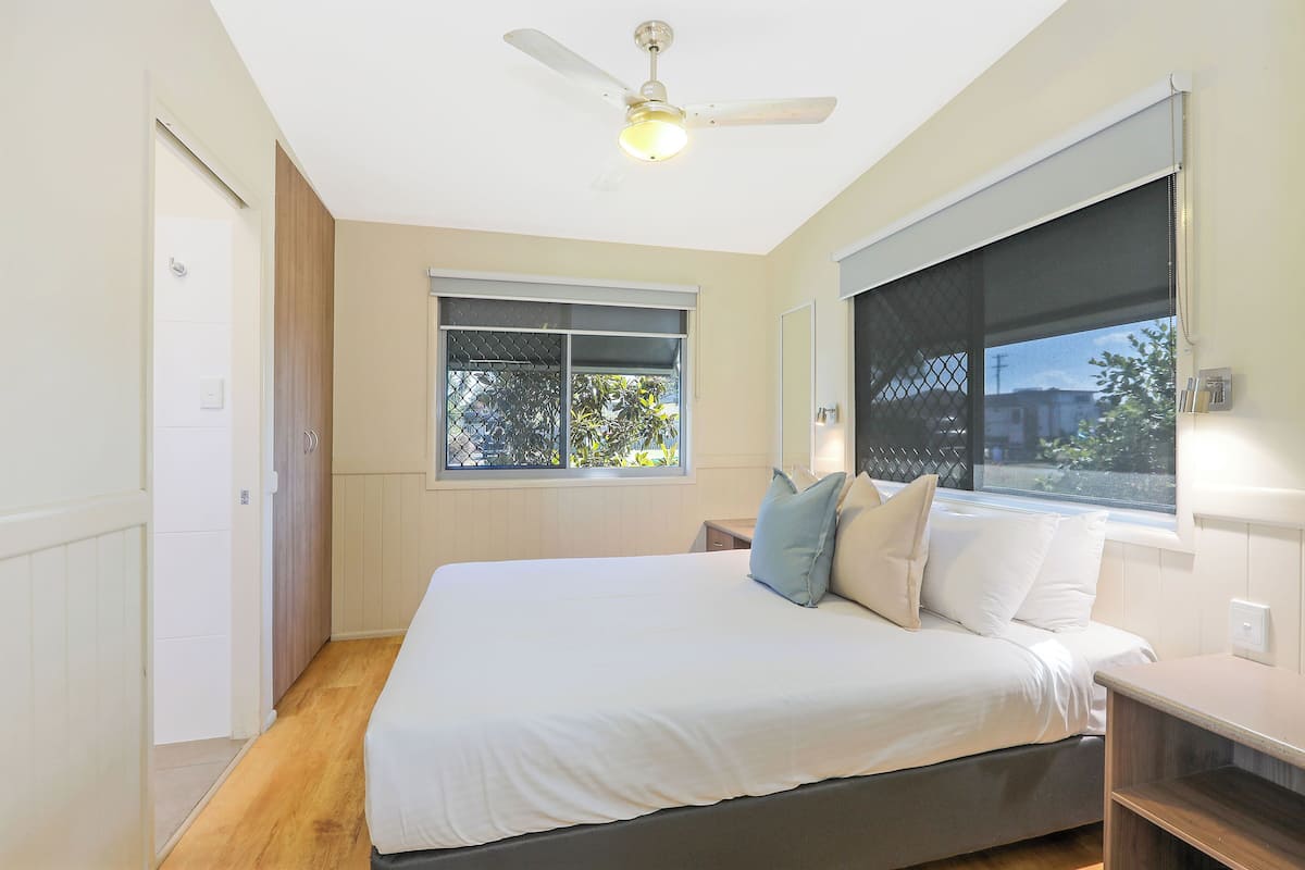 Quarto com cama, ventilador de teto, janela com persianas e mesa de cabeceira.