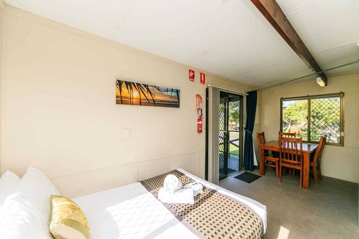 Quarto de hotel com cama, uma mesa de jantar com cadeiras, um extintor de incêndio e vista através de uma porta.
