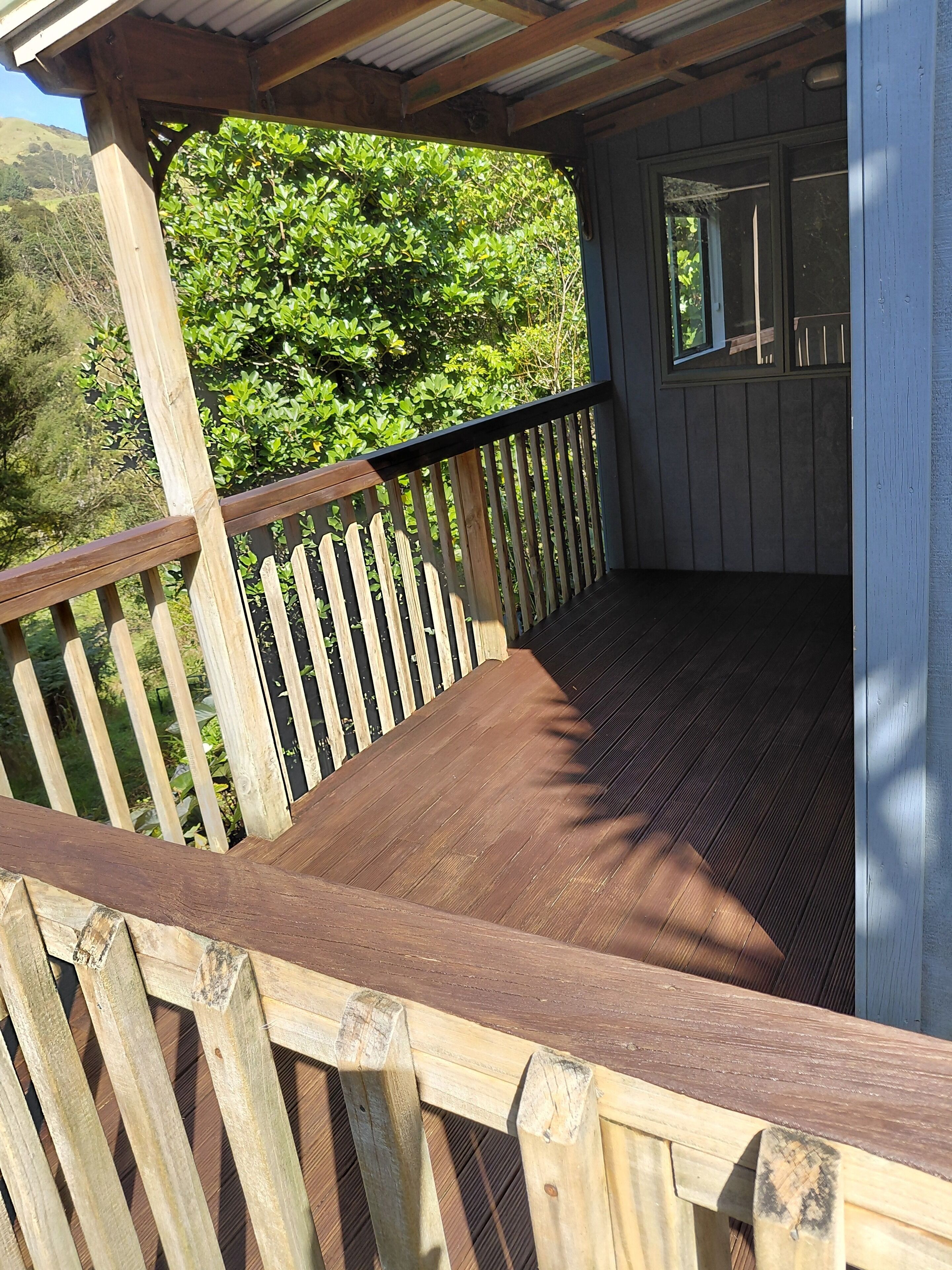 Une terrasse en bois avec une rambarde, un espace abrité doté d’une fenêtre, et une vue sur un feuillage verdoyant en arrière-plan.