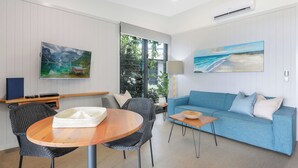 A modern living room with a round wooden table, two black wicker chairs, a blue sofa, and a large window with a view of greenery. - Reflections Shaws Bay - Holiday Park (East Ballina)