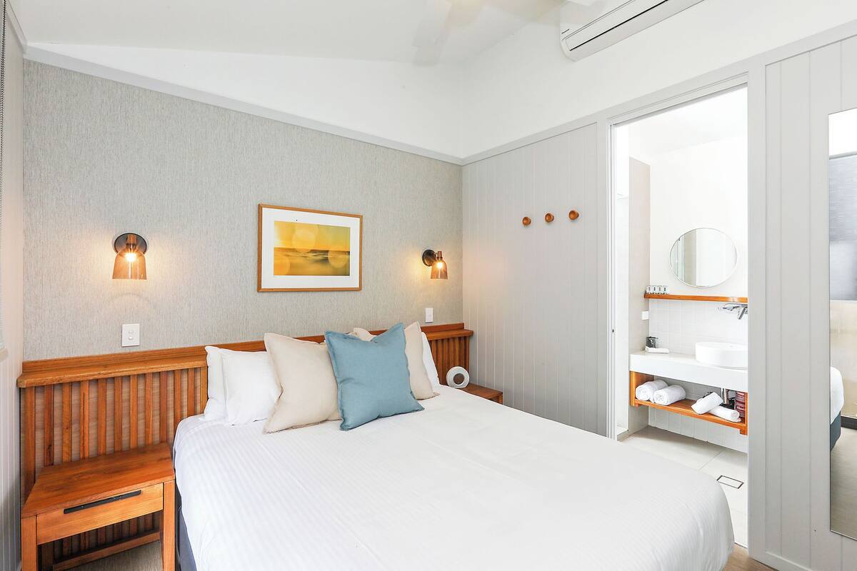 A neatly arranged bedroom with a bed, bedside tables, a wall-mounted light fixture, and a bathroom visible through an open door.