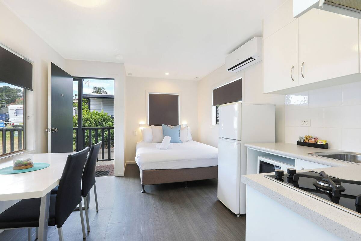 A compact hotel room with a bed, a small kitchenette, a dining area with a table and chairs, and a balcony with a view.