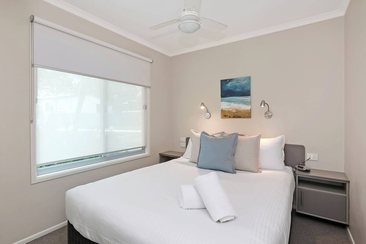 A hotel room with a large bed, a ceiling fan, a painting, and a nightstand with a lamp.