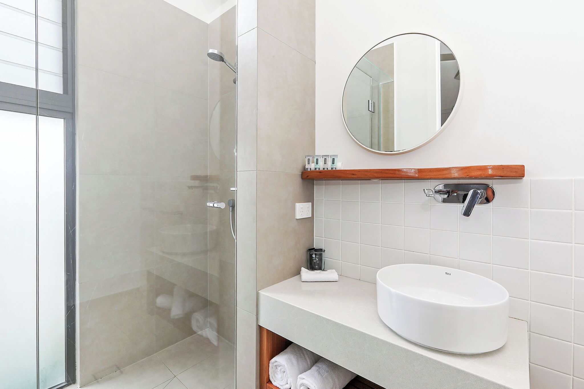 Un bagno moderno con doccia vetrata, uno specchio rotondo, un lavandino bianco e una mensola in legno.