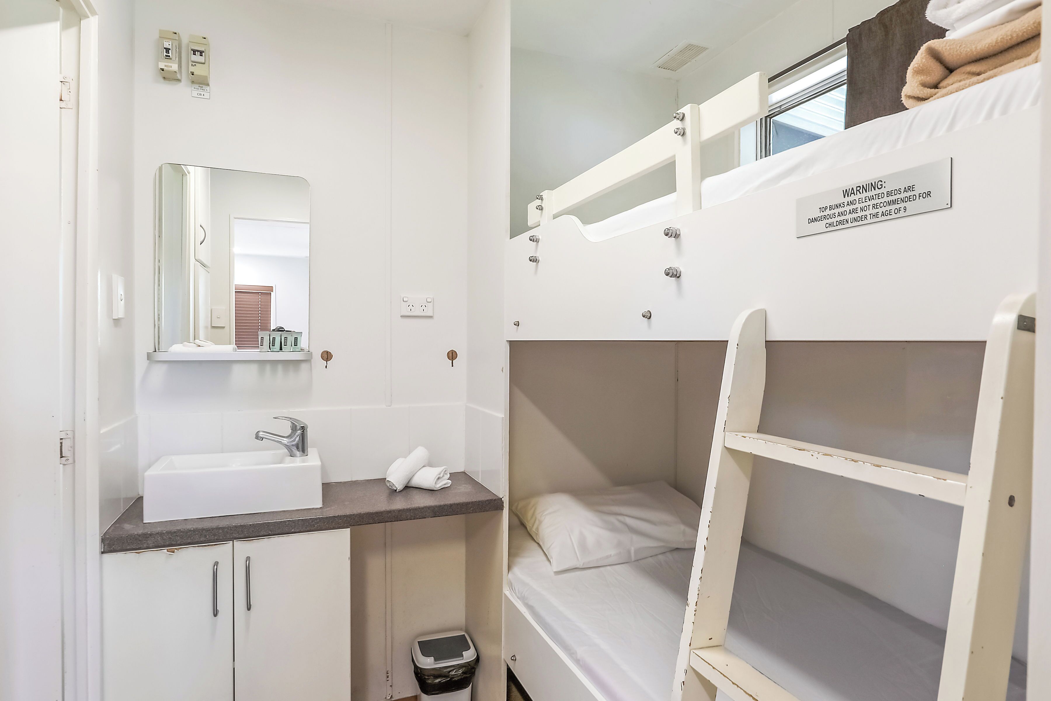A bunk bed with a warning sign, a sink, a mirror, and a small trash bin in a room.