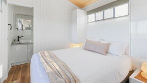 A bedroom with a large bed, a bedside table with a lamp, a window with blinds, and a bathroom with a sink and mirror. - Reflections Shaws Bay - Holiday Park (East Ballina)