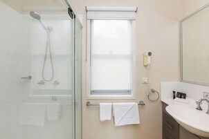 A bathroom with a glass shower enclosure, a window with a blind, a towel rack with two towels, a sink with a mirror, and a hairdryer on the wall.