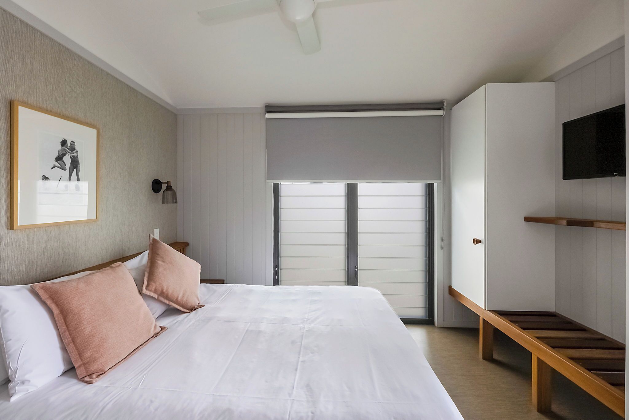 A bedroom with a bed, pillows, a bedside table, a ceiling fan, a framed picture on the wall, a window with blinds, and a TV mounted on the wall.