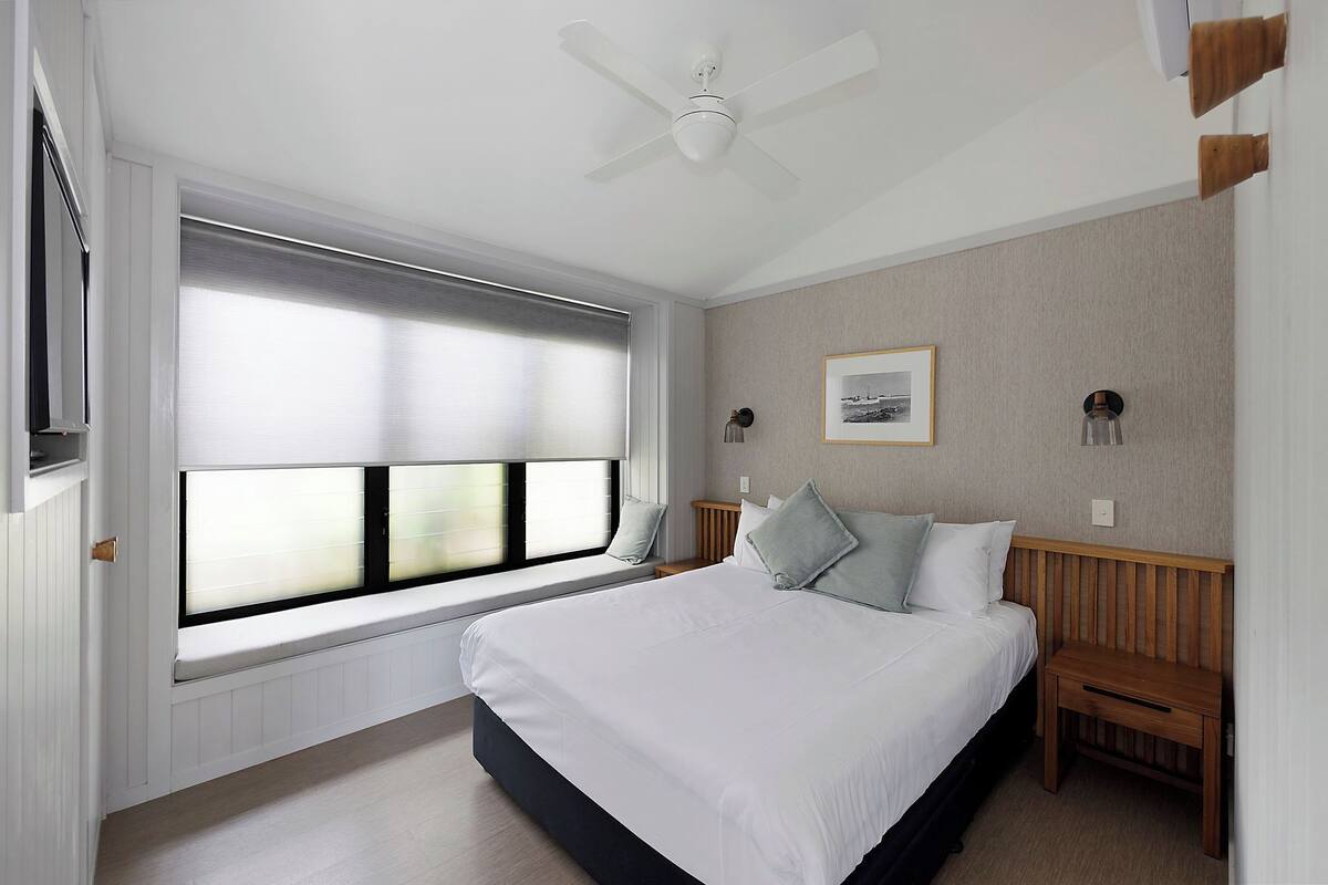 A bedroom with a large bed, a window with blinds, a ceiling fan, and a bedside table.