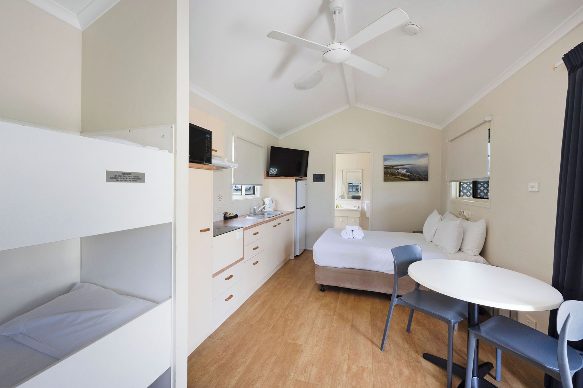 A compact hotel room with bunk beds, a single bed, a small kitchenette, a ceiling fan, a TV, and a window with a view.