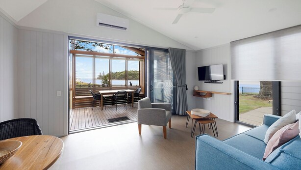 A spacious living room with a large sliding glass door leading to an outdoor deck, a flat-screen TV mounted on the wall, and a cozy seating area with a blue sofa and a small table. - Reflections Evans Head - Holiday Park (Evans Head)