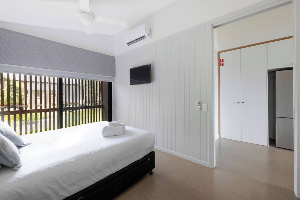A bedroom with a bed, a television mounted on the wall, a door, and a ceiling fan.