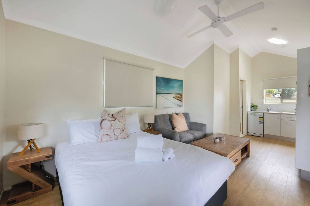 A bedroom with a large bed, a desk, a chair, a ceiling fan, and a view of the kitchen.