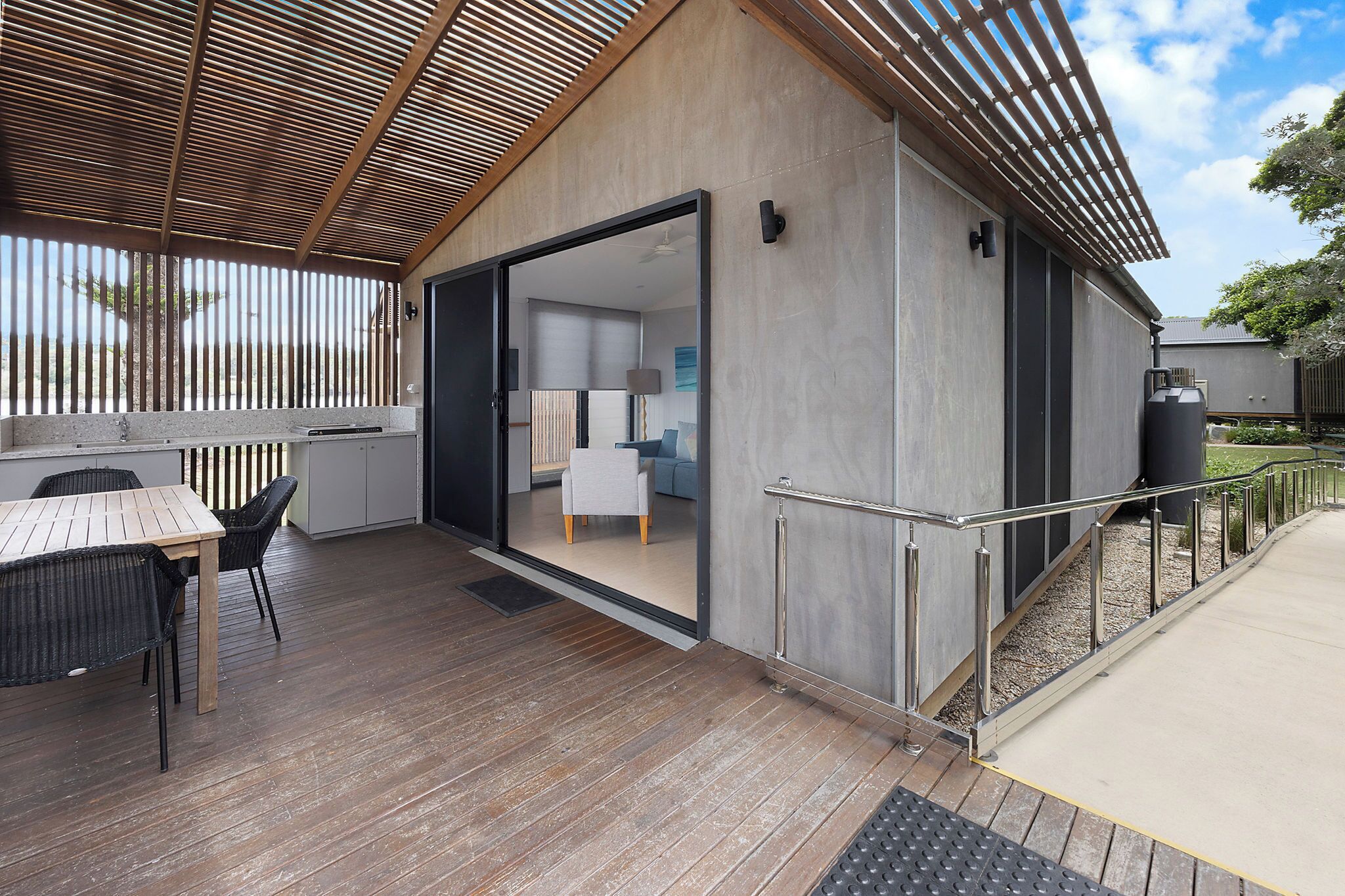 An outdoor patio with a wooden table and chairs, a sliding glass door leading to an interior space with a sofa and lamp, and a concrete deck with a metal railing.