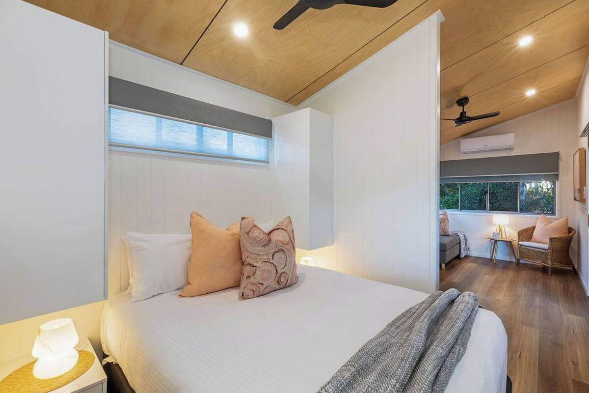 A modern bedroom with a large bed, a bedside table with a lamp, a ceiling fan, and a window with blinds.