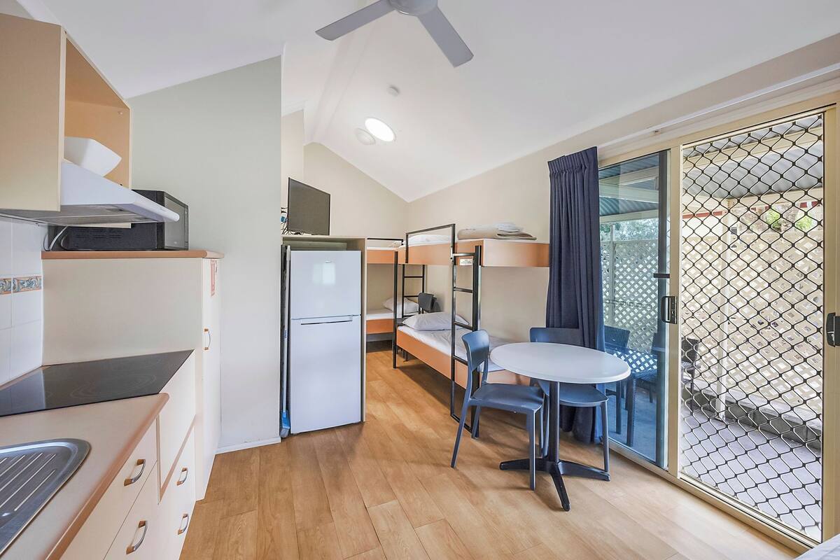 A compact hotel room with a kitchenette, a single bed, a small table with two chairs, and a sliding glass door leading to an outdoor area.
