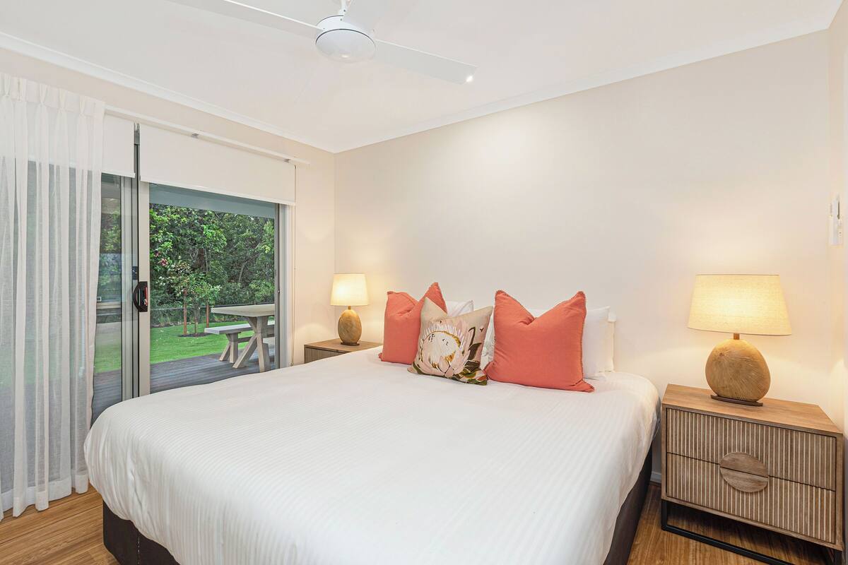 A bedroom with a large bed, two bedside tables with lamps, a sliding glass door to a balcony, and a ceiling fan.