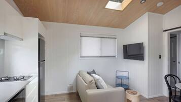 Una cocina moderna con estufa a gas, horno integrado y refrigerador. Hay un sofá blanco con cojines, una mesita con una silla azul y un taburete de madera. Un televisor está montado en la pared sobre un ventanal.