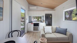 A modern kitchen with white cabinets, a built-in oven, and a refrigerator. There is a dining area with a table and chairs, and a beige sofa with pillows. - Reflections Evans Head - Holiday Park (Evans Head)