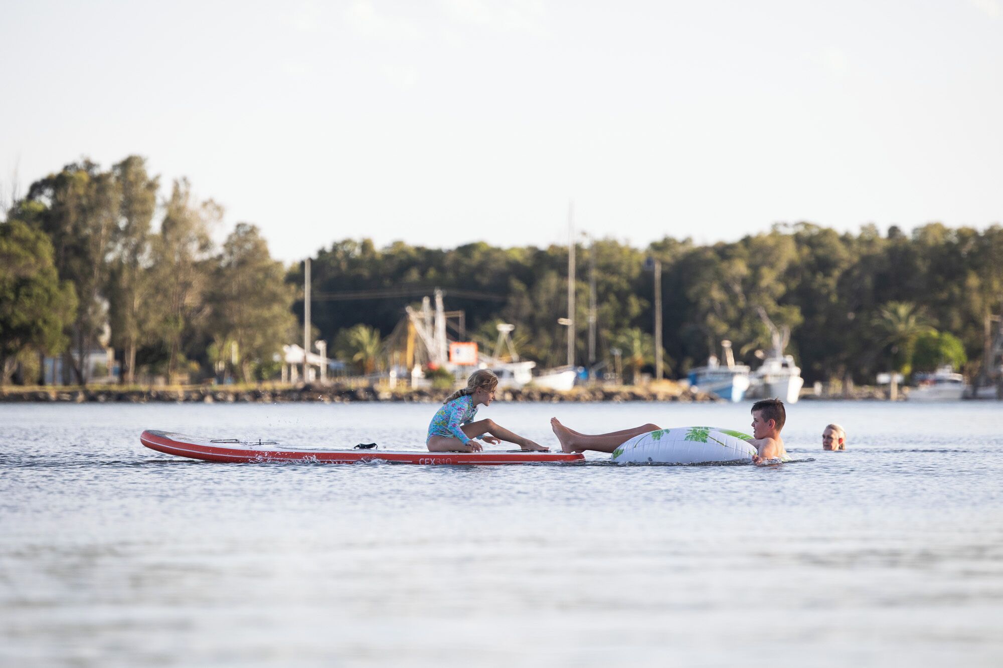 A person lying on a surfboard in calm water with boats and trees in the background.