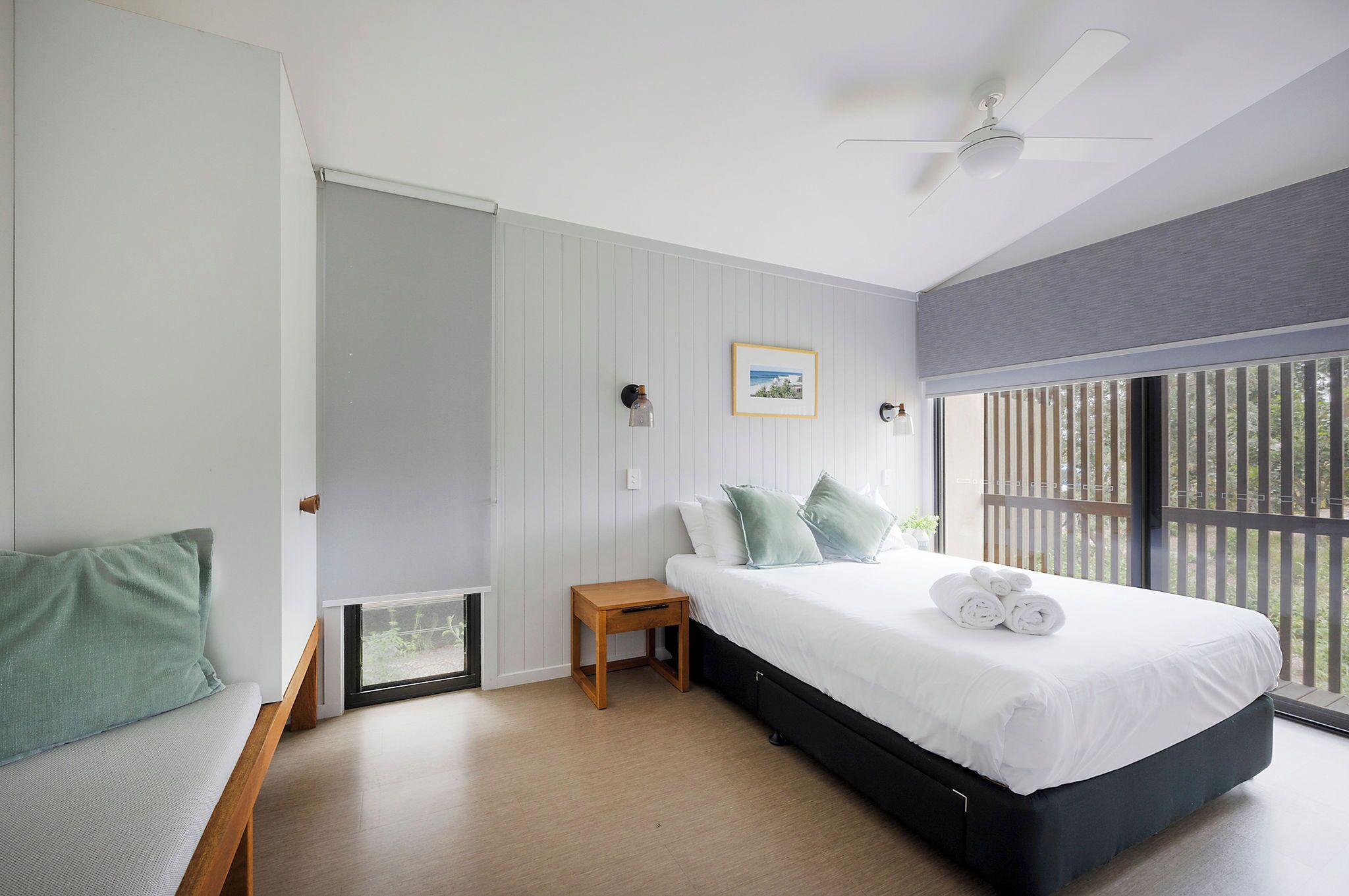A modern bedroom with a large bed, a small wooden nightstand, a bench, and a view of the outdoors through large windows.