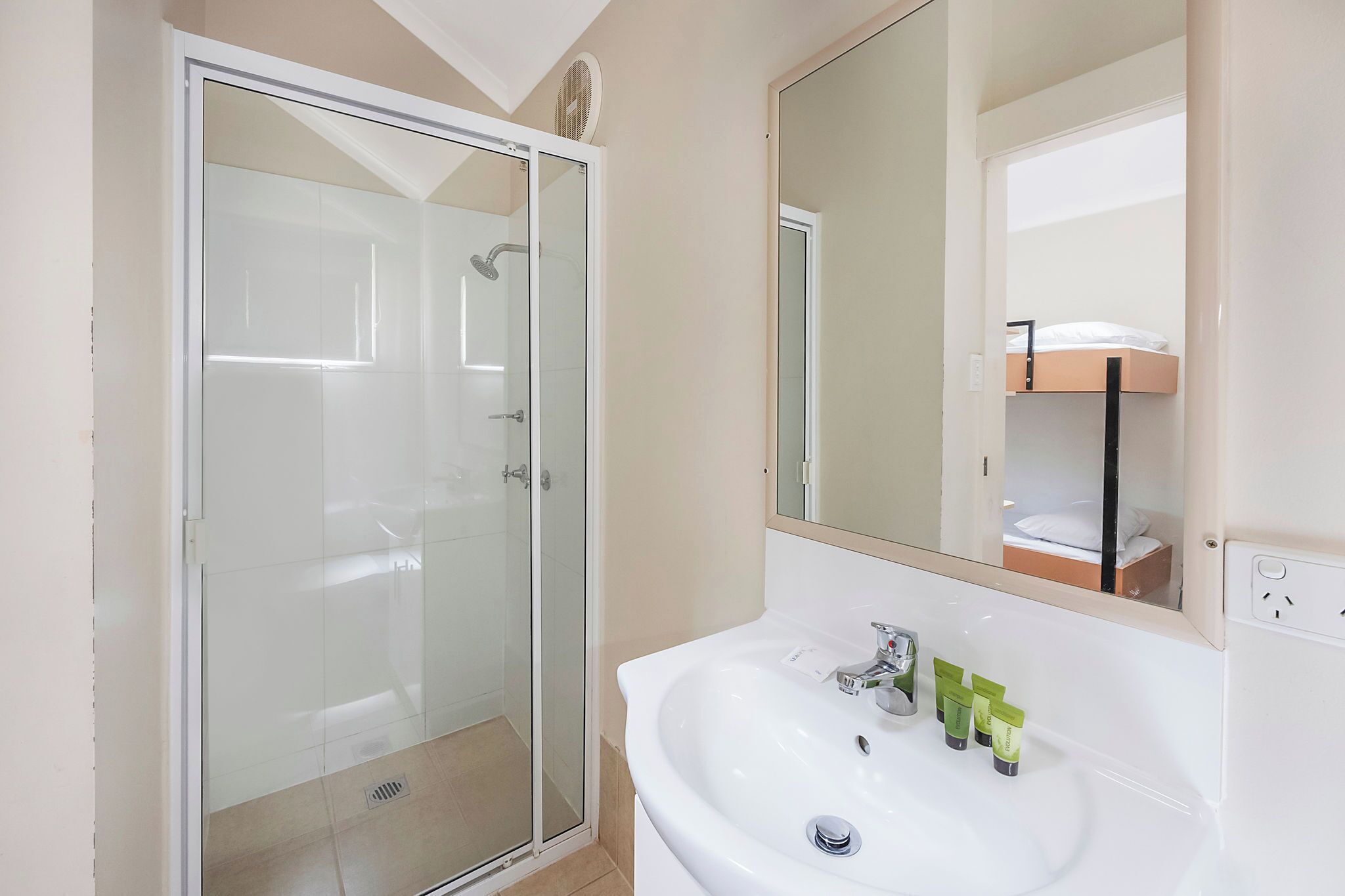 A bathroom with a glass-enclosed shower, a white sink with toiletries, and a mirror reflecting a bunk bed.