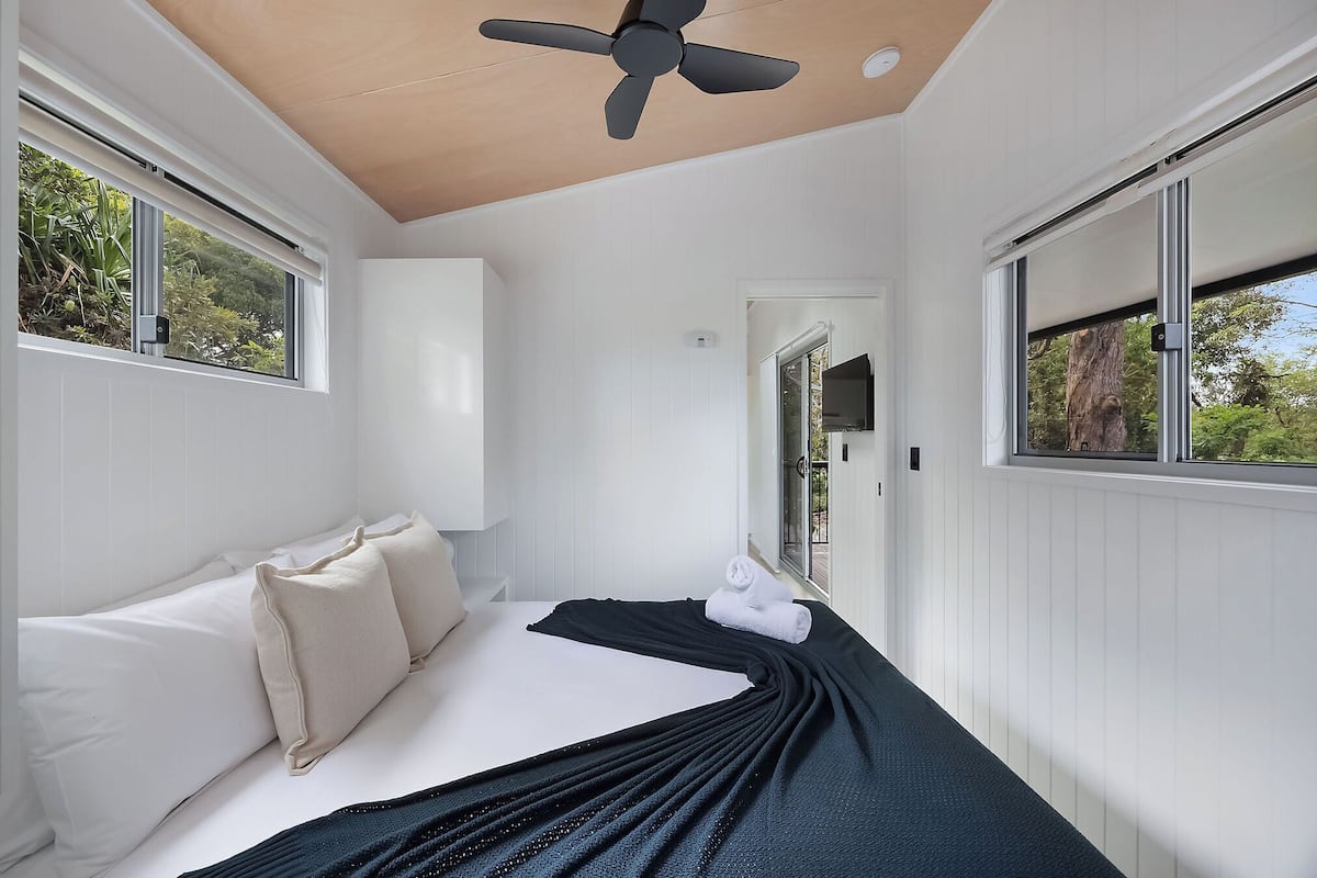 A bedroom with a bed, pillows, a ceiling fan, and windows with views of greenery.